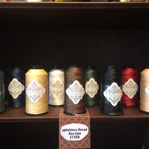 May include: A collection of nine cones of upholstery thread in various colors including grey, black, yellow, white, brown, green, and red. Each cone features a white label with the text Wingback and a decorative floral pattern design.