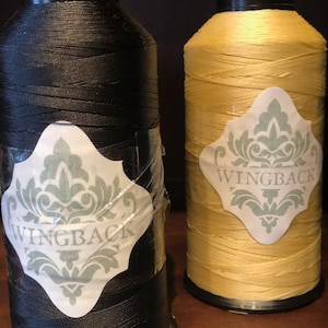 May include: Two large spools of thread, one black and one yellow, each featuring a white diamond-shaped label with a sage green damask pattern and the word WINGBACK printed in capital letters. The black spool has a clear adhesive tape overlay.