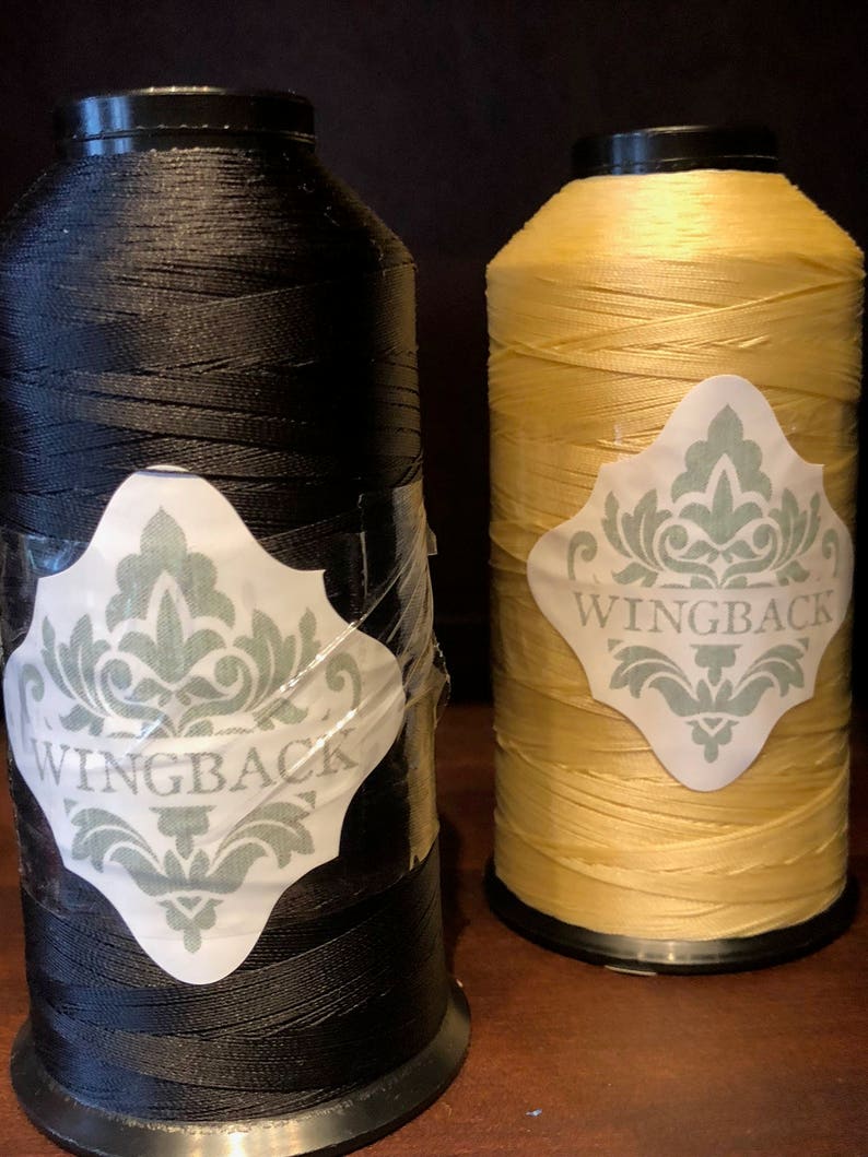 May include: Two large spools of thread, one black and one yellow, each featuring a white diamond-shaped label with a sage green damask pattern and the word WINGBACK printed in capital letters. The black spool has a clear adhesive tape overlay.