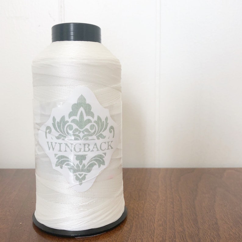 May include: A large spool of white thread with a black cap and base. A white label is attached to the front, featuring a sage green damask pattern and the word WINGBACK printed in capital letters across the center of the decorative design.
