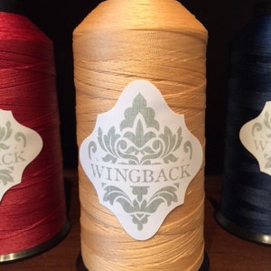 May include: A spool of golden-yellow thread with a black top and bottom cap. A white label is centered on the spool, featuring a light green damask pattern and the word WINGBACK printed in a clear, dark font across the center of the label.