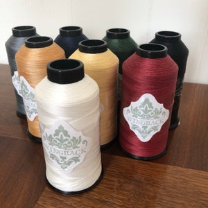 May include: A collection of eight spools of thread in various colors including white, tan, maroon, dark green, navy, grey, and black. Each spool features a black top and bottom with a white label displaying the text WINGBACK in a decorative font.