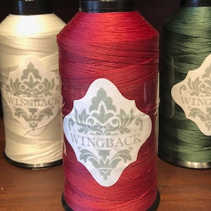 May include: Three large spools of thread in white, deep red, and dark green. Each spool features a white diamond-shaped label with a gray damask pattern and the text WINGBACK printed in the center. The thread is wound tightly around the cylinders.