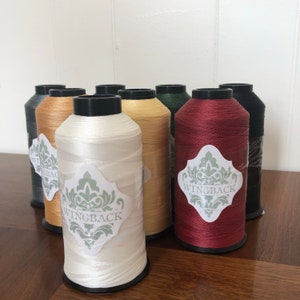 May include: A collection of eight large spools of thread in various colors including white, burgundy, gold, dark green, and black. Each spool features a white label with a green decorative damask pattern and the text WINGBACK printed in black.
