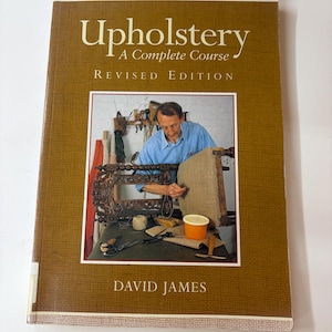 Peut inclure: Un livre à couverture souple intitulé Upholstery A Complete Course Revised Edition par David James. La couverture brune présente une photographie d'une personne travaillant sur un cadre de chaise en bois avec divers outils et fournitures.