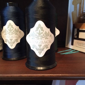 May include: Two large spools of thread, one black and one dark blue, each featuring a white label with a decorative gray damask pattern and the word WINGBACK printed in the center. The spools have black plastic caps and bases.