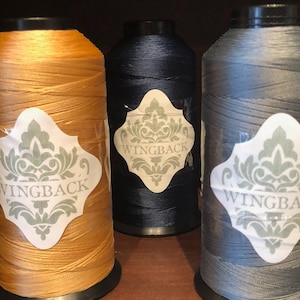 May include: Three large spools of thread in mustard yellow, dark navy blue, and slate gray. Each spool features a white label with a decorative green damask pattern and the text WINGBACK printed in black capital letters on the front.