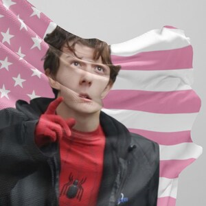 Puede incluir: Una persona con una camiseta roja y negra de Spider-Man y una chaqueta negra, con guantes rojos, se&ntilde;alando. Una bandera rosa y blanca est&aacute; en el fondo.