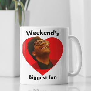 May include: White ceramic mug featuring a red heart with an image of a man wearing sunglasses. The mug has the text "Weekend's Biggest fan" in black. The mug is on a white surface.
