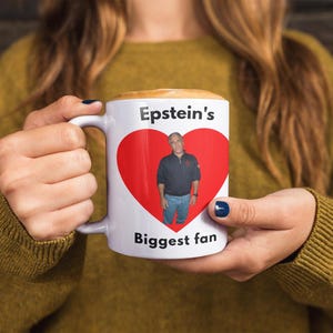 May include: White ceramic mug with the text "Epstein's Biggest fan" and a red heart graphic containing a photo of a man. The mug is held by a person wearing a mustard-coloured jumper.