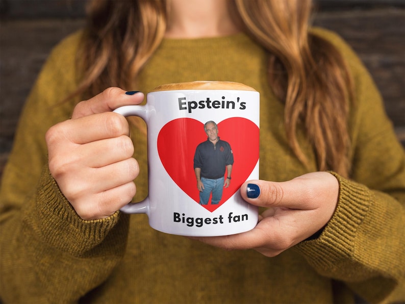 May include: White ceramic mug with the text "Epstein's Biggest fan" and a red heart graphic containing a photo of a man. The mug is held by a person wearing a mustard-coloured jumper.