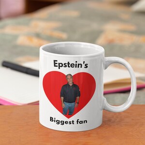May include: White ceramic mug with a black handle. The mug features a red heart with a photo of a man inside. The text "Epstein's Biggest fan" is printed on the mug.