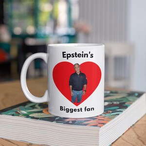 May include: White ceramic mug with a red heart design. The mug has a photo of a man inside the heart, with the text "Epstein's Biggest fan" printed above and below the heart.