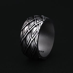 May include: A silver ring with a detailed geometric pattern. The ring has a wide band with a repeating design of pointed shapes. The inside of the ring is smooth and the metal is stamped with "925". The ring is displayed against a dark background.