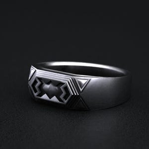 May include: A silver ring with a geometric design. The ring has a rectangular face with a central pattern of interlocking shapes and triangles. The band is smooth and polished, set against a dark background.