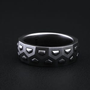 May include: A silver-colored ring with a geometric pattern of half-hexagons and small, raised triangles. The ring has a wide band and a matte finish, suitable for everyday wear. The ring is shown against a dark background.