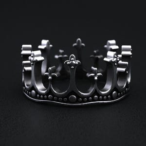 May include: A silver-colored crown ring with a detailed design. The ring features a series of arches and decorative elements, including small spheres and cross-like shapes. The ring is set against a dark background.