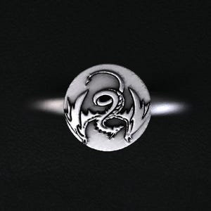 May include: A silver ring with a circular face featuring a detailed dragon design. The dragon is depicted with wings and a curled body. The ring is set against a dark background, highlighting the metallic sheen of the jewelry.