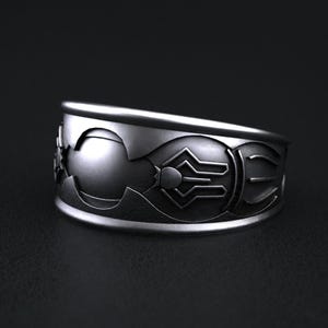 May include: A silver-colored ring with a detailed, engraved design. The ring features a series of stylized shapes and patterns around its band. The design includes a central, rounded element flanked by geometric and abstract motifs. The ring is set against a dark background.