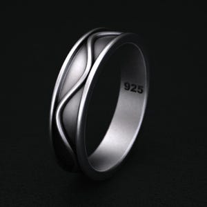 May include: A silver ring with a wave-like design. The ring has a matte finish and the number "925" is stamped on the inside. The ring is set against a dark background.