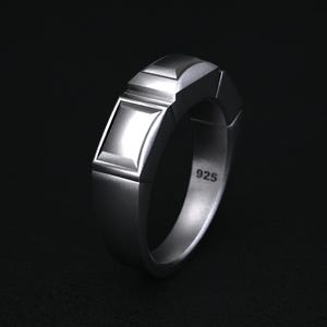 May include: A silver ring with a geometric design. The ring features a rectangular face and a series of angled facets. The number "925" is stamped on the inside of the band. The ring is displayed against a dark background.