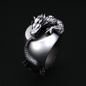 May include: A silver-colored ring featuring a detailed dragon design. The dragon wraps around the ring, with its head resting on the top. The ring has a smooth, polished finish, and the dragon's scales and features are intricately carved.