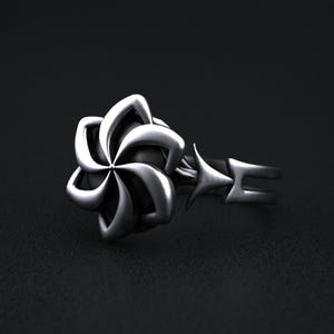 May include: A silver-colored ring featuring a flower design with six petals. The ring has a unique geometric design with star-shaped accents and a split band. The ring is set against a dark background.