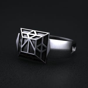 May include: A silver ring with a geometric square design. The square features a complex pattern of lines and shapes, creating a three-dimensional effect. The ring is set against a dark background.