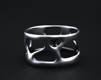 May include: A silver-colored ring with an open, geometric design. The ring has a wide band with irregular cutouts, creating a unique, modern aesthetic. The ring is displayed on a dark surface, highlighting its metallic sheen.