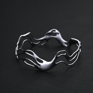 May include: A silver-colored, wavy bangle bracelet. The bracelet has a fluid, organic design with multiple curved lines and a smooth, polished finish. The bracelet is displayed on a dark background.