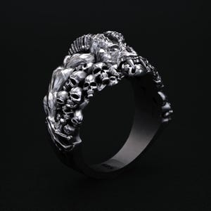 May include: A silver ring featuring a detailed design of skulls and a central figure. The ring has a dark, gothic aesthetic, with intricate carvings and a polished finish. The ring is set against a dark background.