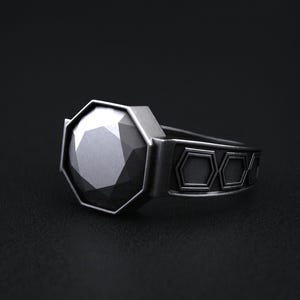 May include: A silver ring with a large, faceted, black gemstone. The ring's band features a repeating geometric design. The gemstone is octagonal and catches the light, creating a sparkling effect. The ring is set against a dark background.