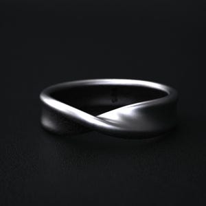 May include: A silver-colored ring with a twisted design. The ring has a smooth, polished surface and a modern, minimalist aesthetic. The ring is set against a dark background, highlighting its shape and metallic sheen. This ring is a piece of jewelry.
