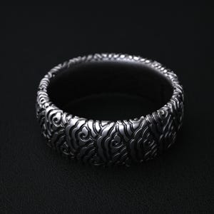 Può includere: Un anello color argento con un intricato motivo ripetuto di disegni a forma di nuvola. L'anello ha una fascia larga e un interno scuro, su uno sfondo nero. Il design dell'anello è dettagliato e strutturato.