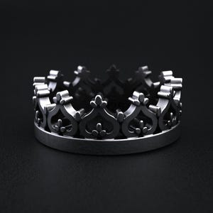 May include: A silver-colored ring in the shape of a crown. The ring features a detailed design with alternating heart and fleur-de-lis shapes. The ring is set against a dark background.