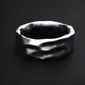 May include: A silver-colored ring with a textured, uneven surface. The ring has a wide band and a dark interior, set against a dark background. The ring's design appears to be organic and handcrafted.