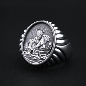 May include: A silver ring featuring an oval medallion with a detailed relief of a man in a boat, holding a fishing net. The ring has a ribbed band design and a polished finish, suitable for religious or nautical themes.