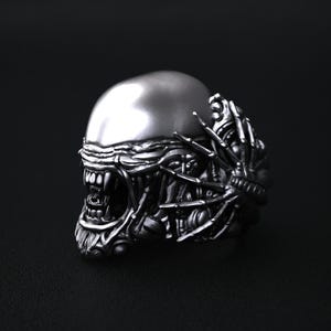 May include: A silver ring with an Alien design. The ring depicts the creature's head, with a detailed skull, sharp teeth, and mechanical elements. The ring is set against a dark background.