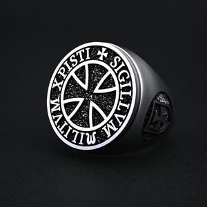 May include: A silver-colored signet ring with a detailed design. The ring features a central cross design within a circular border, with the words "X PISTI SIGILLVM MILITVM" inscribed around it. A smaller cross is visible on the side of the ring.