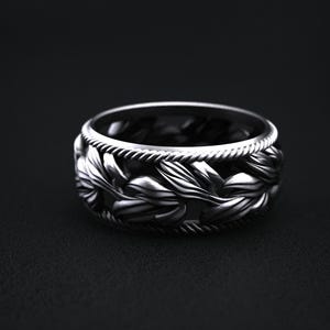 May include: A silver ring with a detailed floral design. The ring features a band of leaves and a rope-like border. The ring is set against a dark background, highlighting its intricate details.