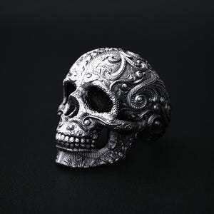 May include: A detailed silver skull ring with intricate floral and scrollwork patterns. The ring's design features a realistic skull shape with open eye sockets and a detailed jawline. The ring is set against a dark background, highlighting its metallic sheen.