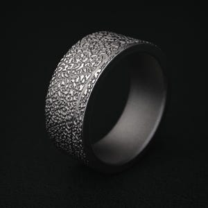 May include: A silver-colored ring with a textured surface. The ring has a wide band with a detailed, irregular pattern. The interior of the ring is smooth and a darker shade of gray. The ring is set against a black background.
