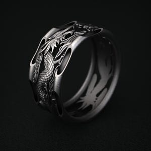 May include: A silver-toned ring with a detailed dragon design. The dragon wraps around the ring, with intricate scales and features. The ring has a dark background, highlighting the metallic sheen and the dragon's form.