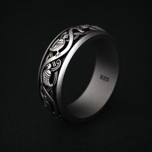 May include: A silver ring with an intricate design featuring birds and interwoven patterns. The ring is marked with "925" on the inside. The ring has a matte finish and is set against a dark background.