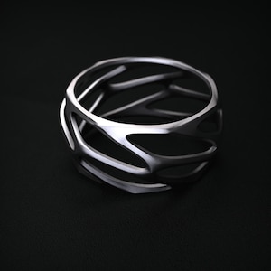 Può includere: Un bracciale a polsino color argento con un design aperto a gabbia. Il bracciale presenta più barre curve e interconnesse che creano un motivo intricato. Il metallo ha una finitura opaca. Il bracciale è appoggiato su una superficie scura.