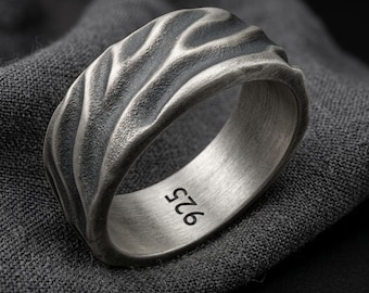 May include: A silver ring with a wide band featuring a textured, wavy, and organic surface pattern. The interior of the band is smooth and clearly stamped with the hallmark 925, indicating the silver content of this metallic jewelry piece.