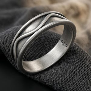 Può includere: Un anello color argento spazzolato caratterizzato da un motivo a onde in rilievo lungo la fascia esterna. L'interno dell'anello è liscio e chiaramente impresso con il numero 925, che indica la composizione metallica del gioiello.