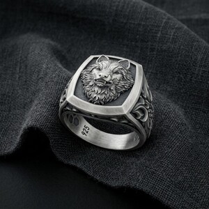May include: A silver-toned ring featuring a detailed relief of a wolf head on the face. The band is engraved with intricate floral patterns along the sides and includes the hallmark 925 stamped on the inner surface of the metal band.
