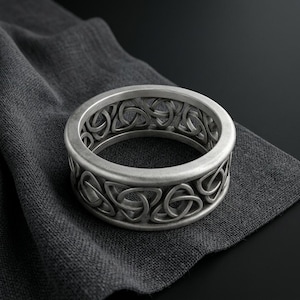 Puede incluir: Un anillo de metal plateado que presenta un diseño de banda calada continua compuesto por patrones repetitivos de nudos triquetra. El anillo tiene un acabado mate cepillado y una estructura de banda ancha y uniforme que muestra los intrincados detalles.