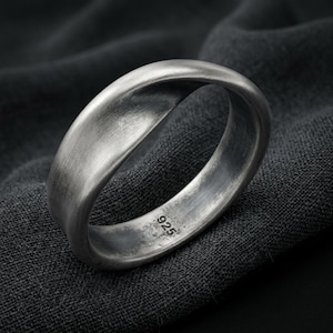 Può includere: Un anello in argento con finitura spazzolata opaca e un design leggermente ritorto. L'interno della fascia presenta il testo inciso 925, che indica la composizione in argento sterling. La superficie metallica mostra una patina grigio scuro.
